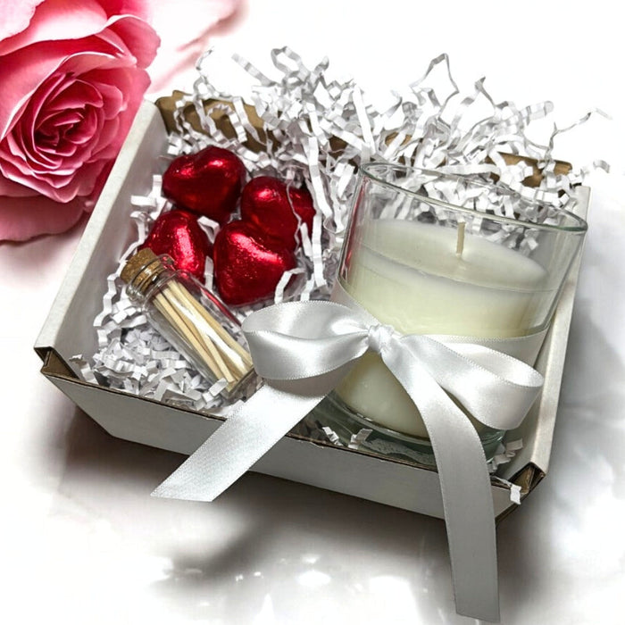 White scented candle in a clear glass jar with a white ribbon, paired with four red foiled heart chocolates and a glass bottle of safety matches.